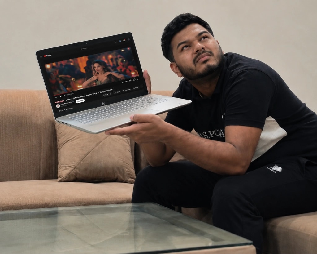 A man sitting on a sofa holds up a laptop playing a YouTube music video and leans towards the screen, straining to hear it because the video is playing too quietly.